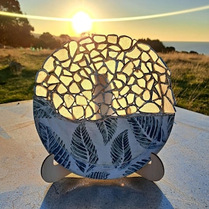 May include: A round, mosaic-style lamp with a leaf-patterned base. The top section is made of irregularly shaped, translucent pieces, allowing light to shine through. The lamp is supported by a wooden stand, and the background features a sunset over a grassy field.