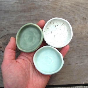 May include: Three small ceramic bowls in green, speckled white, and pale blue. The bowls are held in a hand, highlighting their irregular shapes and textures. These bowls are likely for serving small portions or as decorative items. Measurements are in cm.