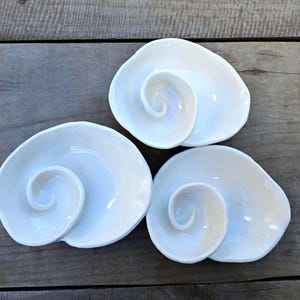 Puede incluir: Tres platos blancos de cerámica con forma de concha. Cada plato tiene un diseño en espiral en el centro, que se asemeja a una concha marina. Los platos están dispuestos sobre una superficie de madera, mostrando su forma y diseño únicos.