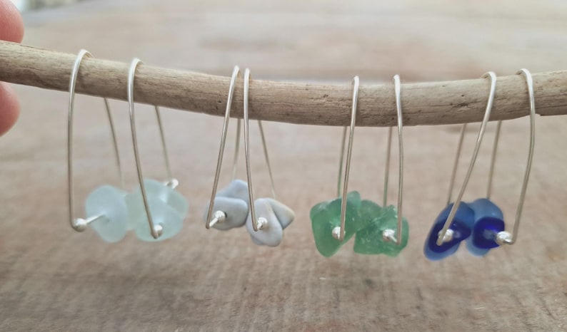 May include: Handmade earrings with sea glass in light blue, grey, green, and blue. Each earring has a silver wire hook and a small silver bead. The earrings are displayed on a wooden stick. The sea glass is in various shapes.