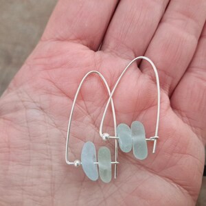 May include: A pair of silver hoop earrings. Each earring has a curved wire design with three light blue, sea glass-style beads. The earrings are presented against a neutral background.