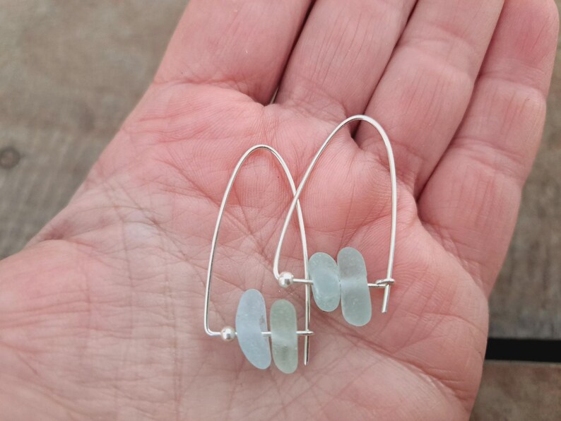 May include: A pair of silver hoop earrings. Each earring has a curved wire design with three light blue, sea glass-style beads. The earrings are presented against a neutral background.