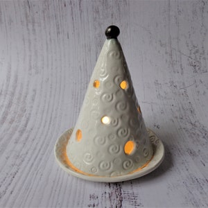 May include: A cream-colored ceramic candle holder shaped like a cone, with a small, dark brown knob at the top. The cone has circular cutouts and a swirling pattern. The candle holder sits on a matching circular base.