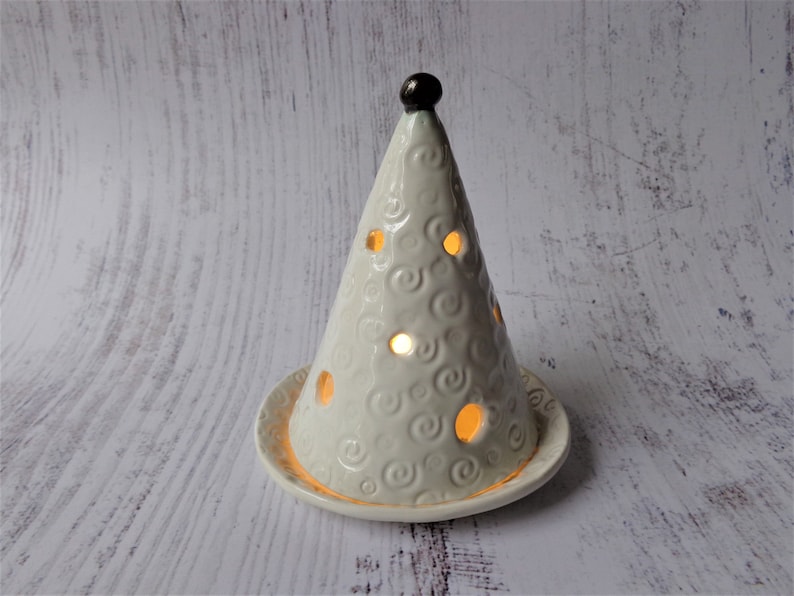 May include: A cream-colored ceramic candle holder shaped like a cone, with a small, dark brown knob at the top. The cone has circular cutouts and a swirling pattern. The candle holder sits on a matching circular base.