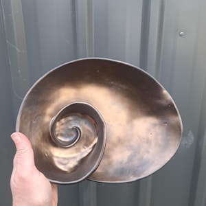 May include: A ceramic bowl with a spiral shell shape, featuring a metallic bronze and dark brown finish. The interior of the bowl has a smooth, curved design that mimics the natural form of a seashell, with a deep central coil and wide rim.