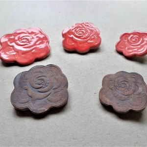May include: Five ceramic rose-shaped decorative pieces. Three are glossy red, and two are matte brown. The rose design is detailed with a central spiral and petal outlines. The pieces vary in size and are arranged on a neutral surface.