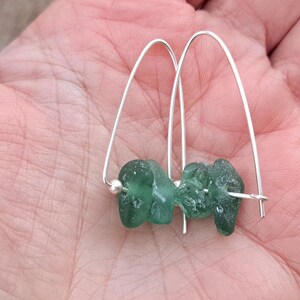 May include: Silver hoop earrings with two pieces of teal green sea glass. The earrings are displayed in a hand, highlighting the natural, organic shapes and the contrast between the metal and glass.