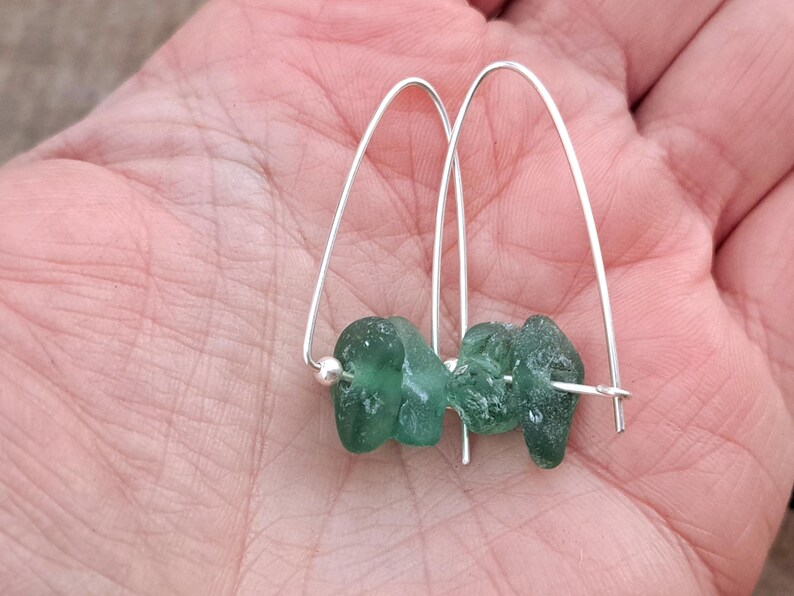 May include: Silver hoop earrings with two pieces of teal green sea glass. The earrings are displayed in a hand, highlighting the natural, organic shapes and the contrast between the metal and glass.