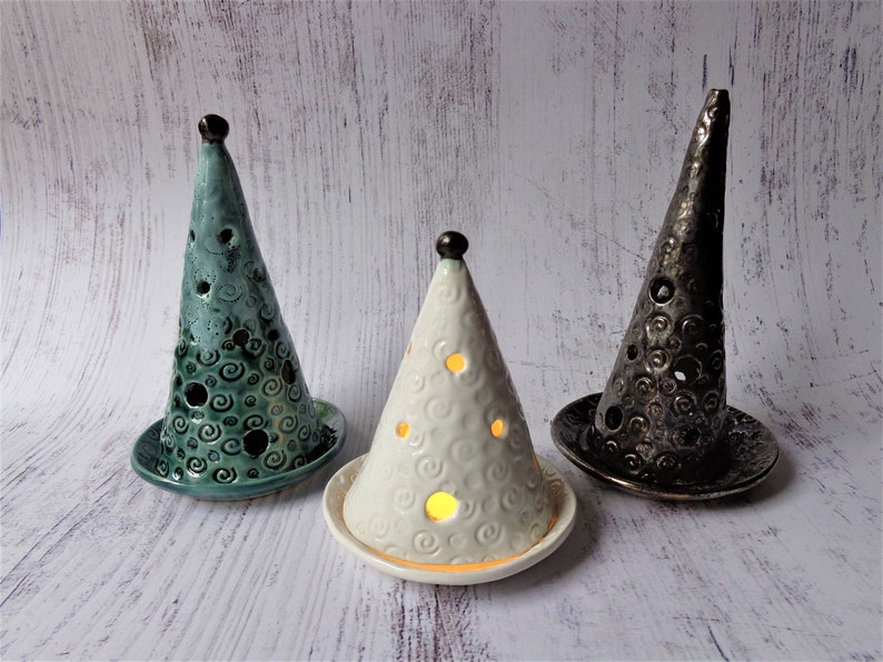 May include: Three ceramic cone-shaped candle holders in teal, white, and black. Each has a circular base and a top finial. The teal and black holders have a swirl pattern and holes. The white holder has a swirl pattern and lit holes.