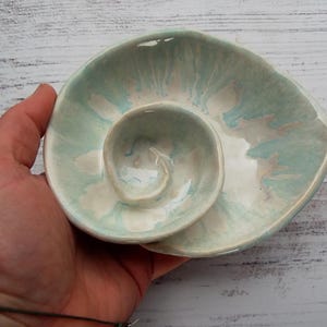 Könnte beinhalten: Eine Keramikschale in Form eines Nautilusgehäuses mit glänzender Oberfläche. Die Oberfläche zeigt ein marmoriertes Muster in sanften Minzgrün-, Hellblau- und Weißtönen. Das Innere hat eine zentrale Spiralstruktur, die sich nach außen wölbt.