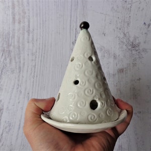 May include: A white ceramic Christmas tree-shaped candle holder. The tree has a conical shape with circular cutouts and a swirl pattern. A small, dark brown ball tops the tree. The candle holder sits on a round base.