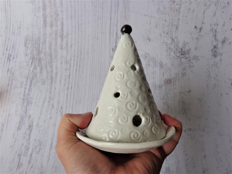 May include: A white ceramic Christmas tree-shaped candle holder. The tree has a conical shape with circular cutouts and a swirl pattern. A small, dark brown ball tops the tree. The candle holder sits on a round base.