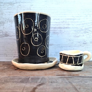 May include: A black ceramic planter with white circular designs and a matching saucer. Beside it is a small black and white ceramic cup with a handle and a matching saucer. The pottery is handmade.