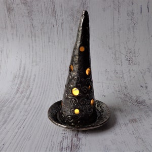May include: A black ceramic candle holder with a conical shape and a circular base. The holder has decorative cutouts that allow light to shine through. The surface has a textured, hammered appearance. The candle holder is designed to hold a small candle.