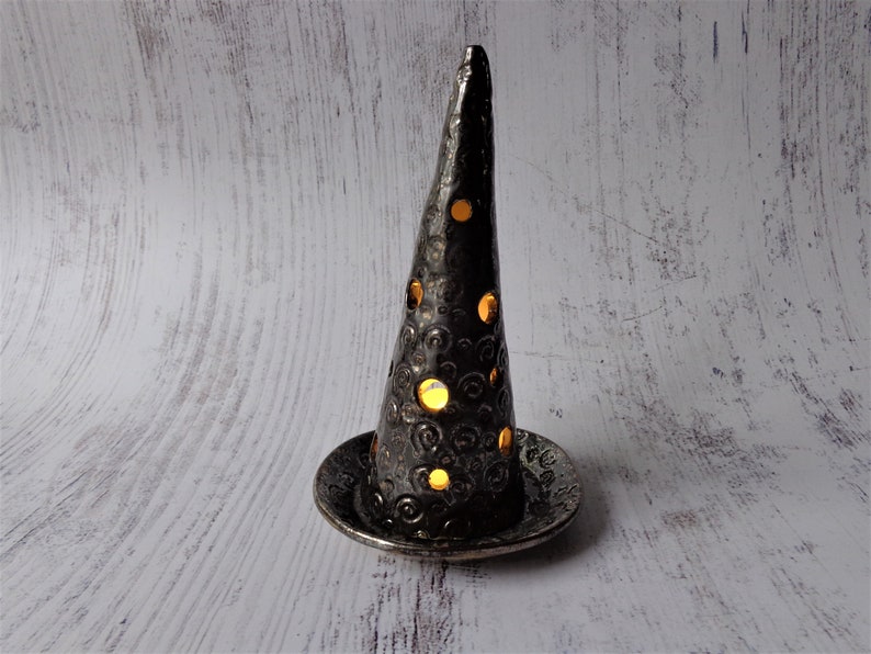 May include: A black ceramic candle holder with a conical shape and a circular base. The holder has decorative cutouts that allow light to shine through. The surface has a textured, hammered appearance. The candle holder is designed to hold a small candle.