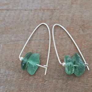May include: Dangle earrings with light green sea glass and silver-coloured wire. The earrings have a curved design with a small silver bead. The sea glass pieces are irregular in shape, offering a natural aesthetic.
