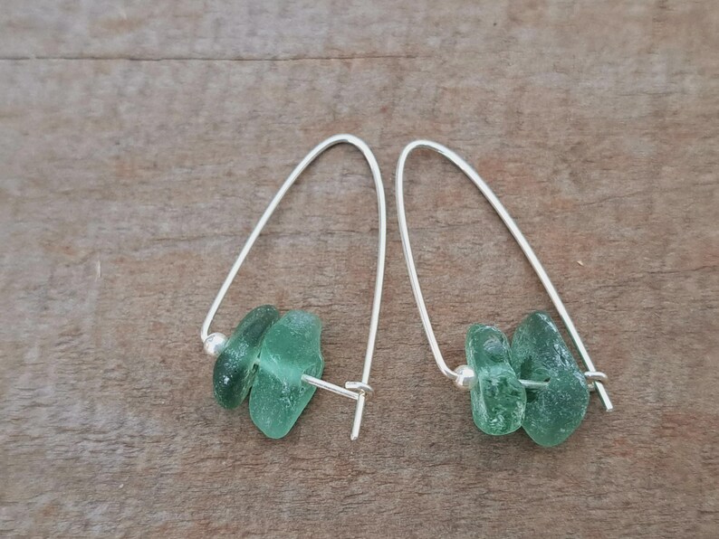 May include: Dangle earrings with light green sea glass and silver-coloured wire. The earrings have a curved design with a small silver bead. The sea glass pieces are irregular in shape, offering a natural aesthetic.