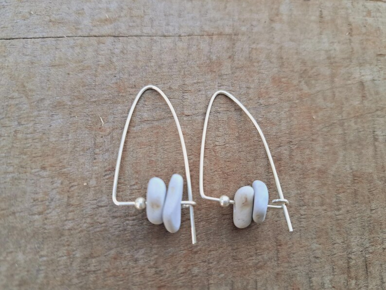 May include: A pair of silver drop earrings. Each earring has a curved wire design, a small, round bead, and three pale blue, oblong beads. The earrings are presented on a wooden surface.