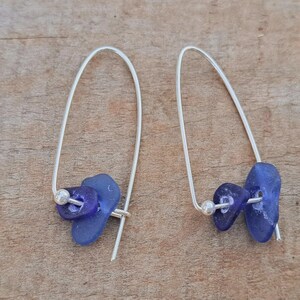 May include: Handmade earrings with blue sea glass and silver-tone metal. Each earring has a curved wire design, two pieces of blue sea glass, and a small silver bead. The earrings are presented on a wooden surface.