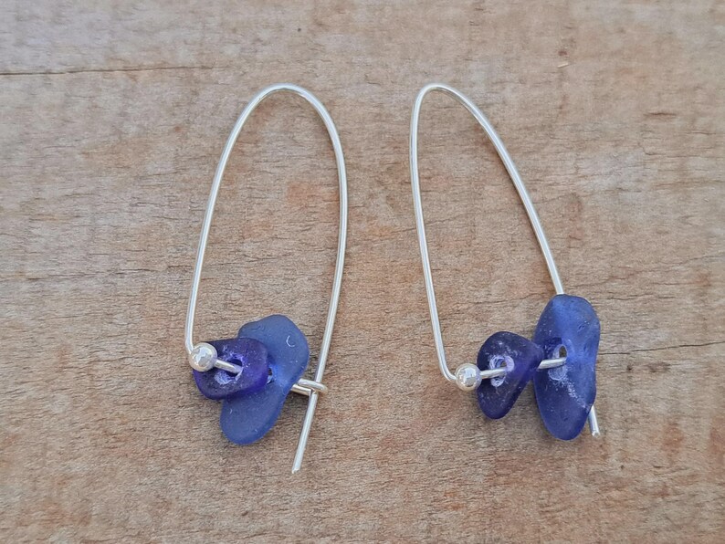 May include: Handmade earrings with blue sea glass and silver-tone metal. Each earring has a curved wire design, two pieces of blue sea glass, and a small silver bead. The earrings are presented on a wooden surface.