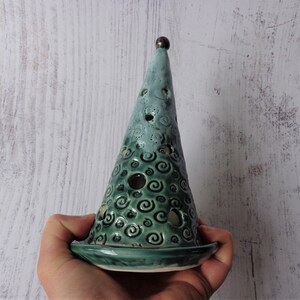 May include: A teal ceramic candle holder shaped like a cone with a flat base. The cone has a series of holes and a swirling pattern. A small, dark brown sphere sits atop the cone. The candle holder is held in a person's hand.
