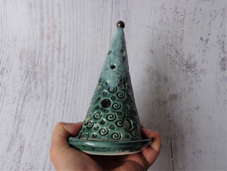 May include: A teal ceramic candle holder shaped like a cone with a flat base. The cone has a series of holes and a swirling pattern. A small, dark brown sphere sits atop the cone. The candle holder is held in a person's hand.