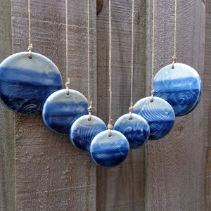 May include: A handcrafted wind chime featuring seven ceramic discs in varying sizes. Each disc is painted with a blue and white wave pattern, suspended by twine. The wind chime hangs against a weathered wooden fence.