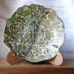 May include: Decorative plate with a unique design. One half features a mosaic of green sea glass pieces, while the other has a white background with a green leaf pattern. The plate is supported by a wooden stand.