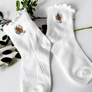 May include: A pair of white ribbed socks with a ruffled top edge. Each sock features an embroidered bee design in yellow, black, and brown. The socks are displayed on a white surface with green foliage.