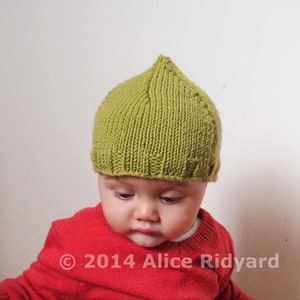 Peut inclure: Un bébé portant un bonnet en tricot vert et un pull rouge. Le bonnet a un sommet pointu et est fait avec un motif de point simple. Le pull est un modèle à col rond.