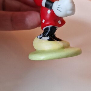 May include: A ceramic figurine of Minnie Mouse wearing a red dress with white polka dots and white gloves. She is standing on a green base.