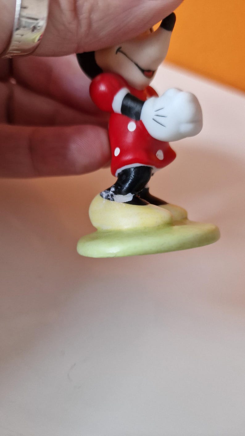 May include: A ceramic figurine of Minnie Mouse wearing a red dress with white polka dots and white gloves. She is standing on a green base.