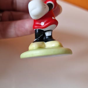 May include: A small ceramic figurine of Minnie Mouse wearing a red dress with white polka dots and a red bow. She is standing on a green base.