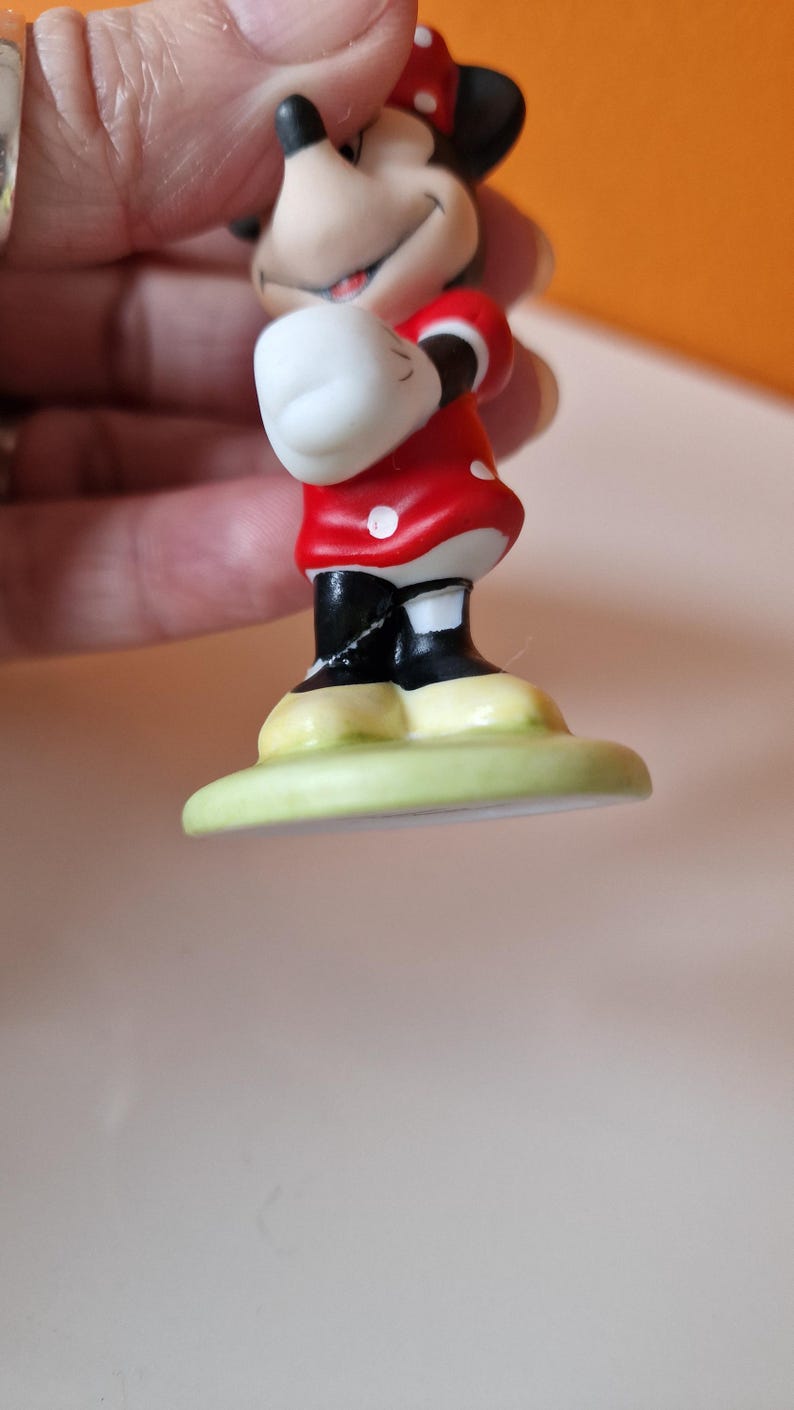 May include: A small ceramic figurine of Minnie Mouse wearing a red dress with white polka dots and a red bow. She is standing on a green base.