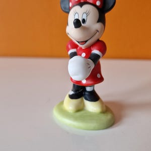 May include: A small ceramic figurine of Minnie Mouse wearing a red dress with white polka dots and a red bow with white polka dots. She is standing on a green base.