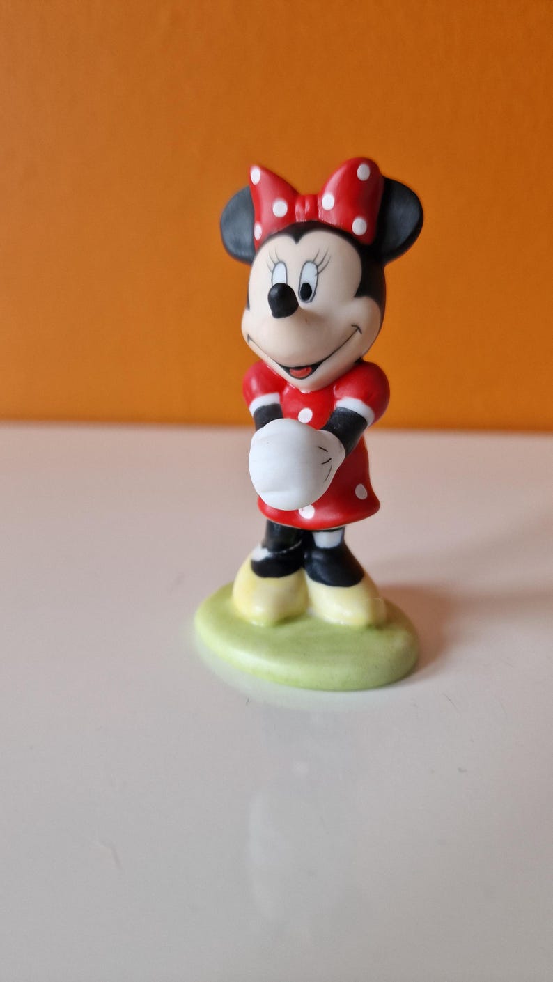May include: A small ceramic figurine of Minnie Mouse wearing a red dress with white polka dots and a red bow with white polka dots. She is standing on a green base.