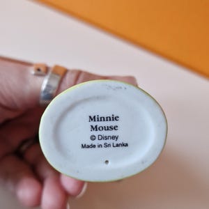 May include: A white ceramic figurine of Minnie Mouse with a green base. The base has the text "Minnie Mouse &copy; Disney Made in Sri Lanka".