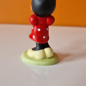 May include: A small figurine of Minnie Mouse, standing on a green base. She is wearing a red dress with white polka dots and a black bow.