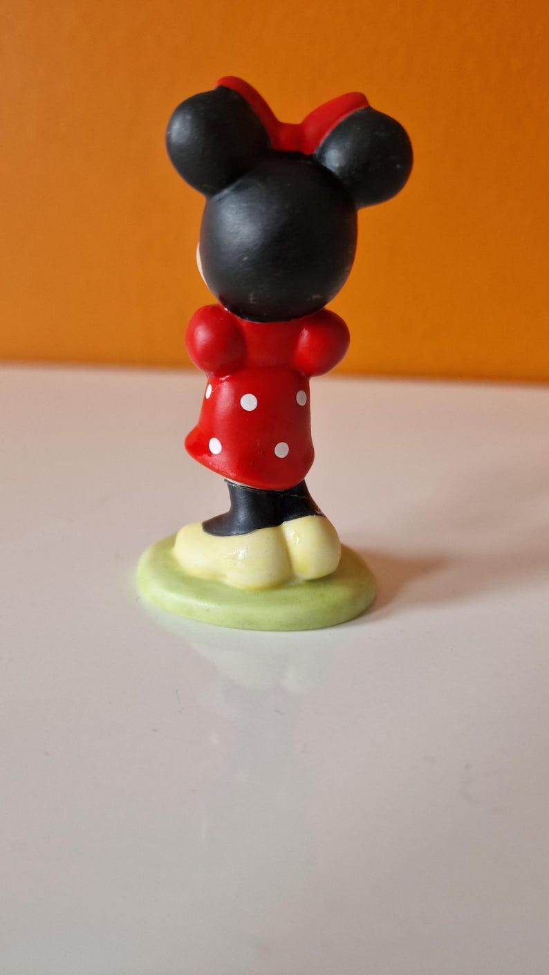 May include: A small figurine of Minnie Mouse, standing on a green base. She is wearing a red dress with white polka dots and a black bow.