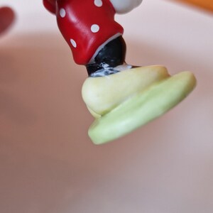 May include: A ceramic figurine of Mickey Mouse standing on a green base. Mickey is wearing his signature red shorts with white polka dots, black shoes, and white gloves.