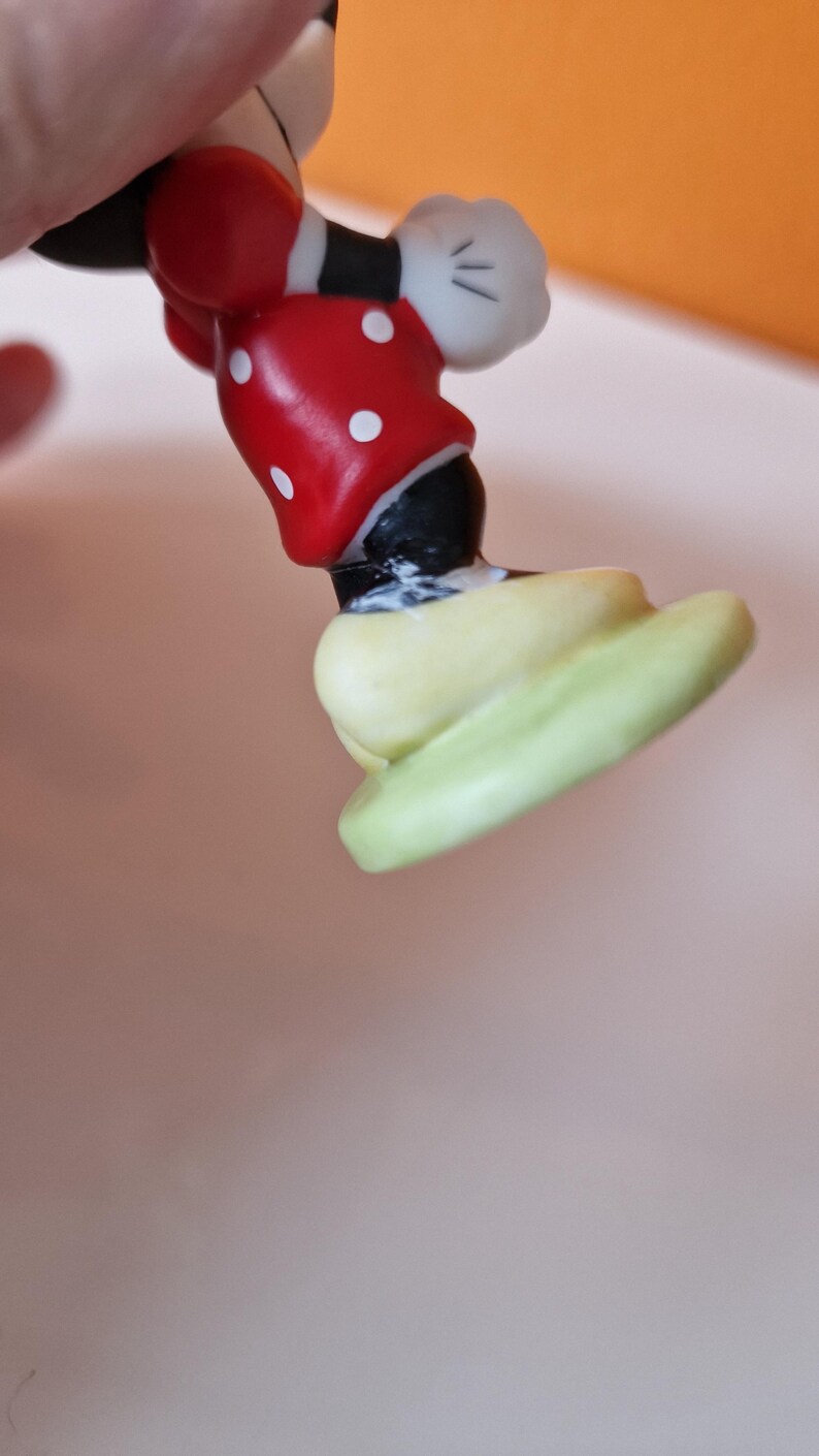 May include: A ceramic figurine of Mickey Mouse standing on a green base. Mickey is wearing his signature red shorts with white polka dots, black shoes, and white gloves.