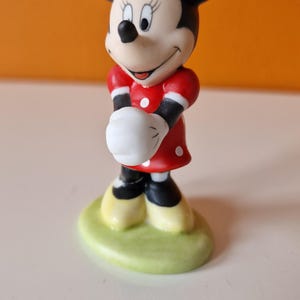 May include: A ceramic figurine of Minnie Mouse wearing a red dress with white polka dots and a red bow with white polka dots. She is standing on a green base.