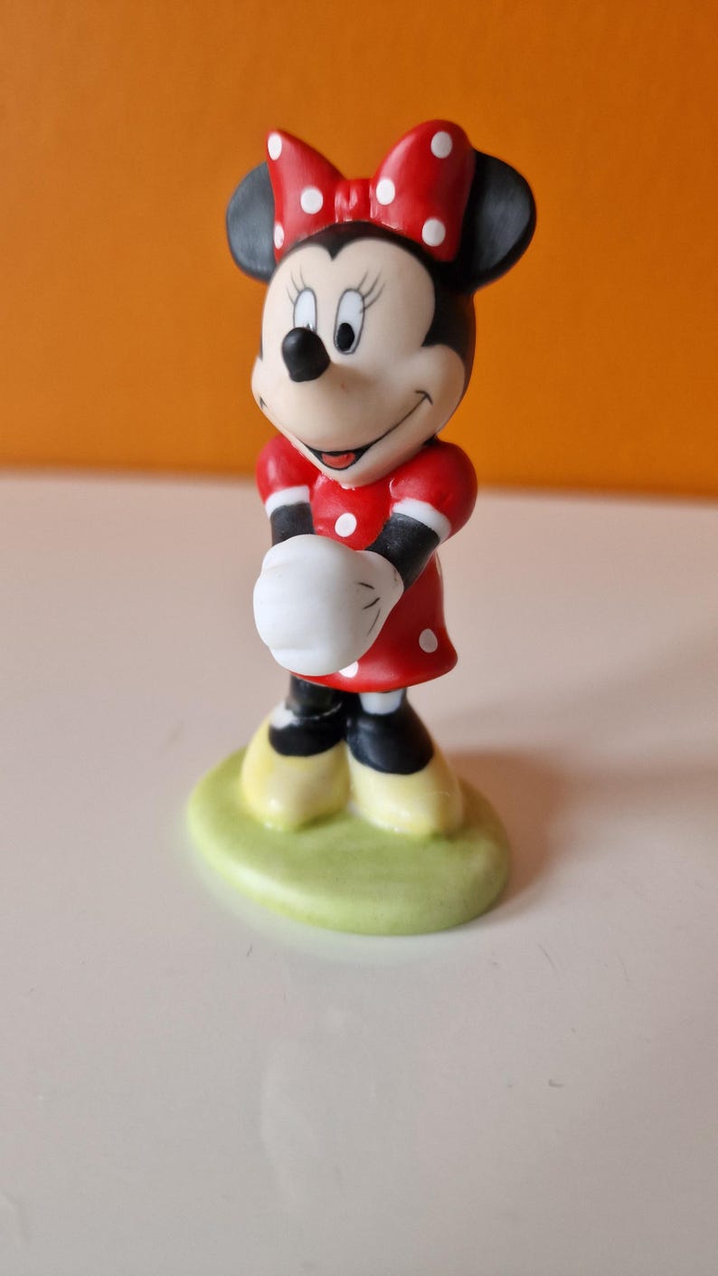 May include: A ceramic figurine of Minnie Mouse wearing a red dress with white polka dots and a red bow with white polka dots. She is standing on a green base.