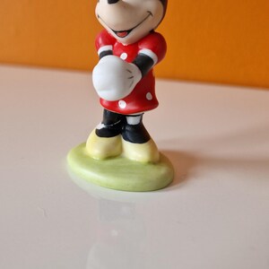 May include: A small ceramic figurine of Minnie Mouse. She is wearing a red dress with white polka dots and a red bow with white polka dots. She is standing on a green base.