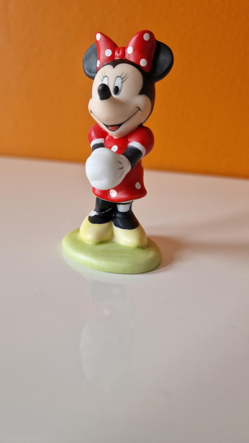 May include: A small ceramic figurine of Minnie Mouse. She is wearing a red dress with white polka dots and a red bow with white polka dots. She is standing on a green base.