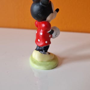 May include: A small ceramic figurine of Minnie Mouse wearing a red dress with white polka dots and a black bow. She is standing on a green base.