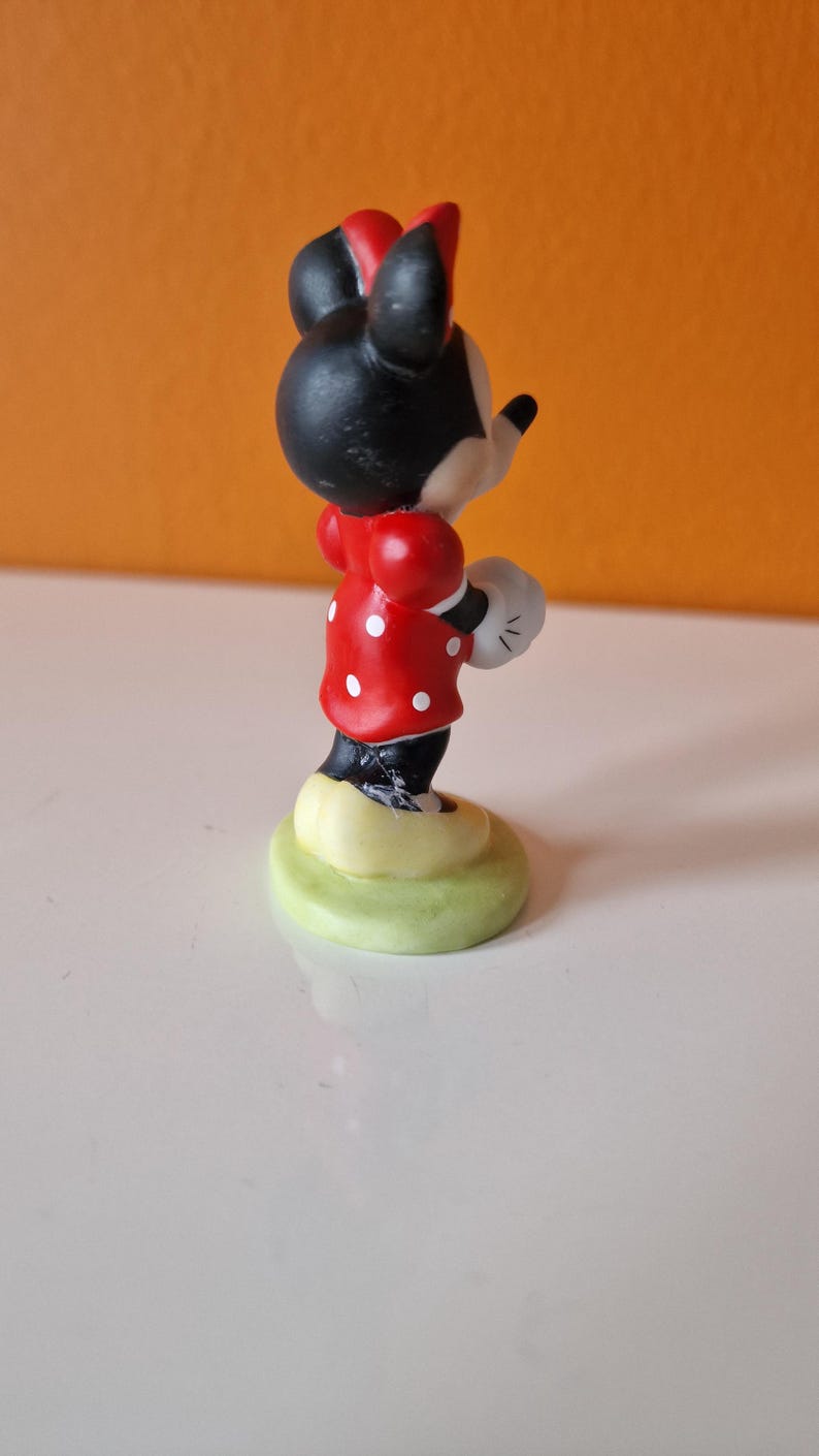 May include: A small ceramic figurine of Minnie Mouse wearing a red dress with white polka dots and a black bow. She is standing on a green base.