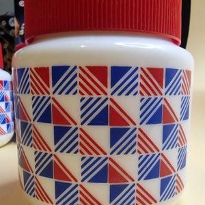 May include: White container with a red, ribbed lid. The body of the container is white with a repeating pattern of red and blue squares and diagonal stripes. The container is cylindrical and appears to be made of plastic.