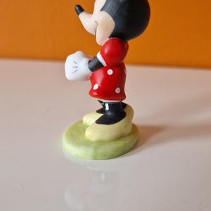 May include: A small figurine of Minnie Mouse standing on a green base. She is wearing a red dress with white polka dots, white gloves, and a red bow.