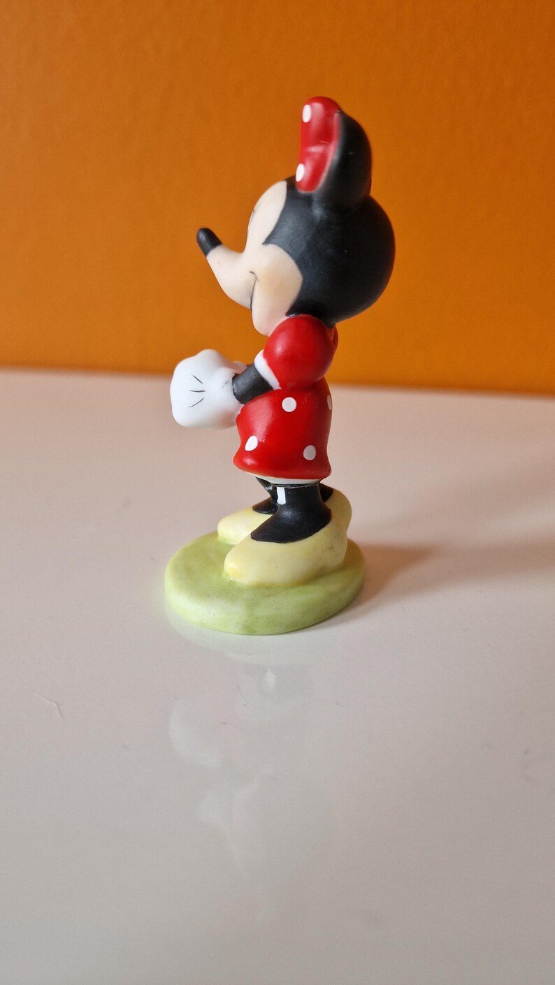 May include: A small figurine of Minnie Mouse standing on a green base. She is wearing a red dress with white polka dots, white gloves, and a red bow.